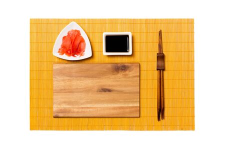 Empty rectangular brown wooden plate with chopsticks for sushi, ginger and soy sauce on yellow bamboo mat background. Top view with copy space for you design.の写真素材