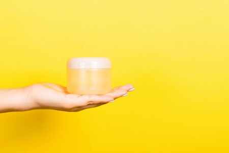 Female hand holding cream bottle of lotion isolated. Girl give jar cosmetic products on yellow...
