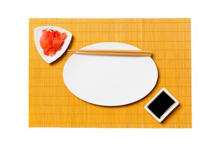 Empty oval white plate with chopsticks for sushi and soy sauce, ginger on yellow bamboo mat background. Top view with copy space for you design.の写真素材