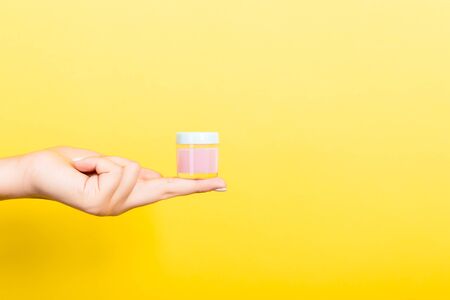 Female hand holding cream bottle of lotion isolated. Girl give jar cosmetic products on yellow...