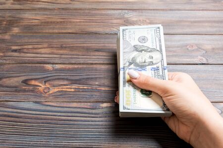 Female hand holding a bundle of one hundred dollar banknotes on wooden background. Salary and wage...