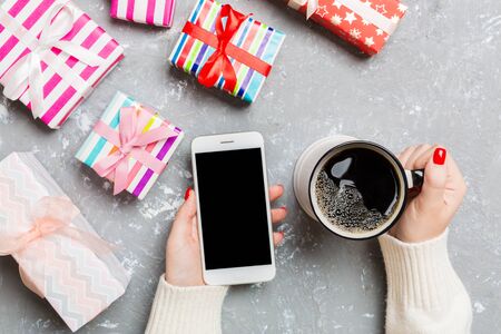 Top view of a woman holding a cup of coffee in one hand and a phone in another hand on cement background. Gift boxes and Christmas decorations. New Year holiday concept. Mockup.の写真素材