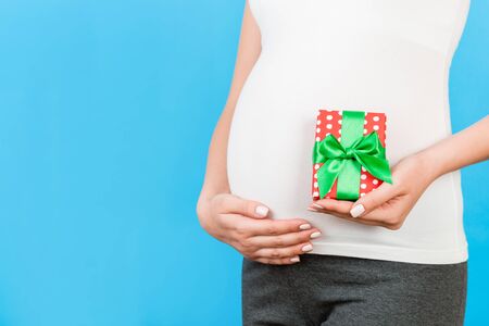 Close up of pregnant woman in home clothing holding gift box at blue background. Baby surprise concept. Happy pregnancy. Copy space.の写真素材