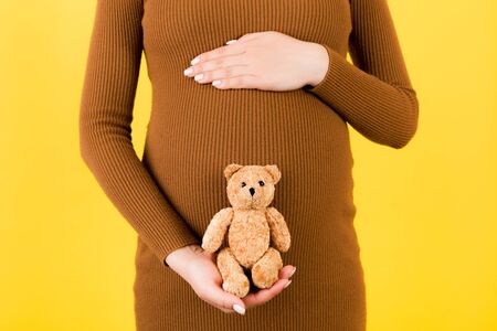 Cropped image of happy pregnant woman in brown dress holding teddy bear against her belly at yellow background. Child expecting. Copy space.の写真素材