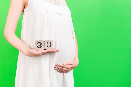 Cropped image of pregnant woman in white dress showing thirty weeks of pregnancy cubes at green...