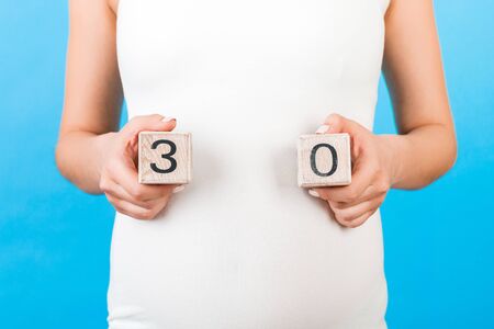 Cropped portrait of pregnant woman holding cubes which show thirty weeks of pregnancy at blue background. Healthy pregnancy. Copy space.の写真素材