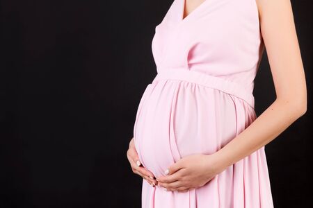 Cropped image of pregnant woman hugging her abdomen. Mother is wearing pink dress at black background. Future parent is expecting her child. Copy space.の写真素材