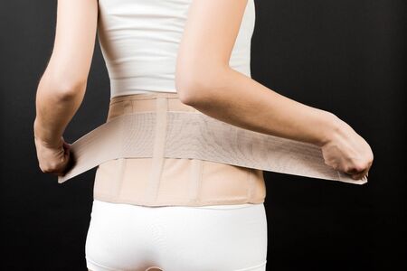 Back view of pregnant woman in underwear putting on supporting bandage to reduce backache at black...