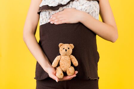Close up of pregnant woman in brown pajamas holding teddy bear against her abdomen at yellow background. Parenthood concept. Copy space.の写真素材