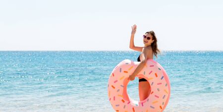 Full length portrait of attractive woman holding doughnut inflatable ring and waving her hand on the beach at the sea background. Have happy summer.の写真素材