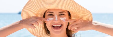Beautiful young woman in hat is applying sunblock under her eyes and on her nose like Indian. Sun...