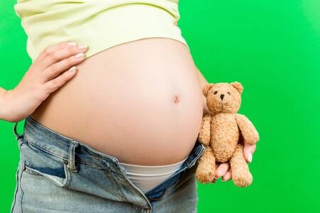 Close up of pregnant woman in unzipped jeans embracing teddy bear near her belly at colorful...
