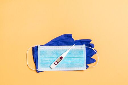 Top view of medical gloves, digital thermometer and surgical mask on orange background with copy...