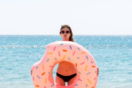 Beautiful woman is playing with donut rubber ring and giving a kiss on the beach at the sea background.の写真素材