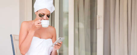Wrapped in towel after shower woman with eye patches is drinking tea or coffe at the table and reading news or messages at the hotel terrace. Girl is enjoying her vacation.の写真素材
