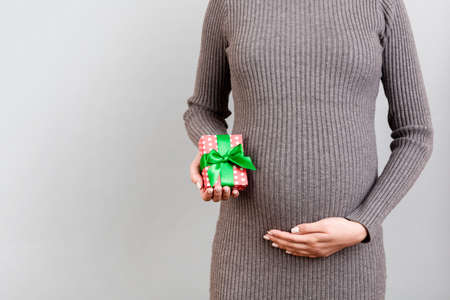 Close up of pregnant woman in gray dress holding gift box at gray background. Baby surprise concept....