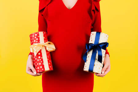 Cropped image of pregnant woman in red dress holding two gift boxes at yellow background. Is it a boy or a girl Waiting for twins. Pregnancy celebration. Copy space.の写真素材