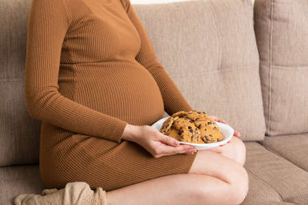 Close up of pregnant woman enjoys eating cookies resting on the sofa at home. Unhealthy sweet pastry during pregnancy concept.の写真素材