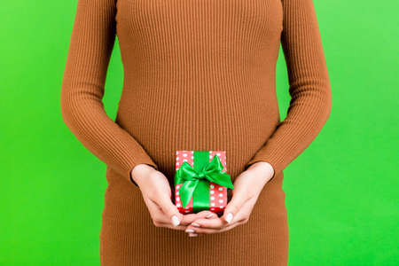 Close up of gift box in pregnant woman's hands against her abdomen at green background. Young mother in brown dress. Happy pregnancy. Copy space.の写真素材