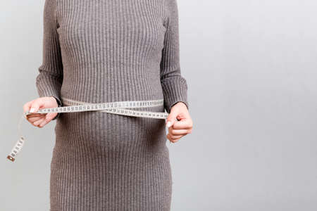 Close up of pregnant woman in gray dress measuring her growing tummy with a tape measure at gray...
