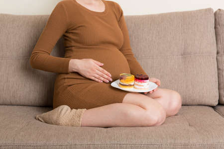 Close up of hungry pregnant woman is eating tasty desserts sitting on the sofa at home. Enjoying sweet food during pregnancy concept.の写真素材