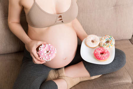 Close up of pregnant woman enjoys eating different donuts resting on the sofa. Unhealthy desserts during pregnancy concept.の写真素材