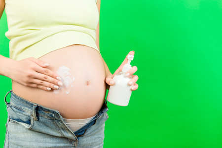 Close up of pregnant woman in opened jeans applying moisturizing lotion on her belly to avoid...