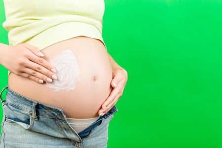 Close up of pregnant woman in opened jeans applying skin care cream on her belly to prevent stretch...