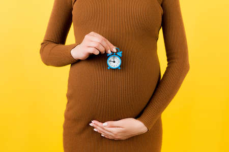 Close up of pregnant woman in brown dress holding alarm clock against her belly at yellow...