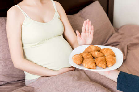 Close up of pregnant woman relaxing in bed makes stop gesture to croissants. Expecting mother refuses to eat pastry. Diet during pregnancy concept.の写真素材