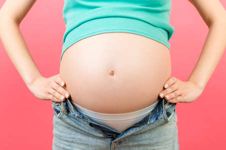 Cropped image of pregnant belly at colorful background with copy space. Expecting mother in opened jeans. Motherhood concept.の写真素材