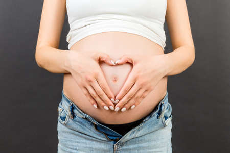 Close up of pregnant woman in opened jeans making heart on her belly at colorful background with copy space. Love to a future baby concept.の写真素材