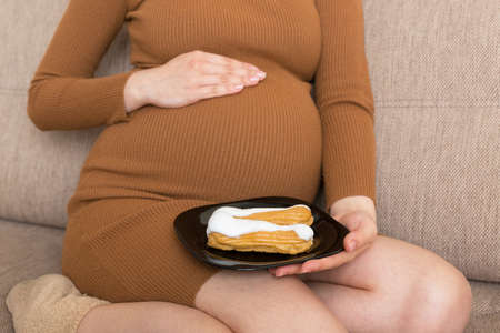Close up of hungry pregnant woman is eating pastry with great pleasure relaxing on the sofa at home. Happy pregnancy concept.の写真素材
