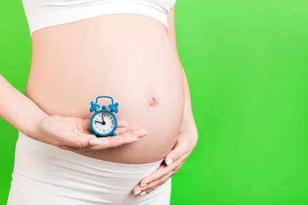 Cropped image of pregnant woman in white underwear holding alarm clock against her abdomen at green...