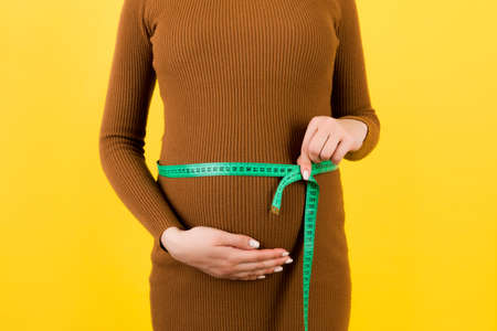 Close up of tape measure on pregnant woman's belly in brown dress at yellow background. Measuring of growing abdomen. Copy space.の写真素材