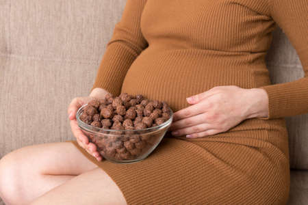 Close up of pregnant woman enjoys eating crunchy chocolate cereal balls from a bowl relaxing on the sofa. Dieting during pregnancy concept.の写真素材