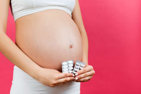 Close up of pregnant woman holding maternal supplements at colorful background with copy space....