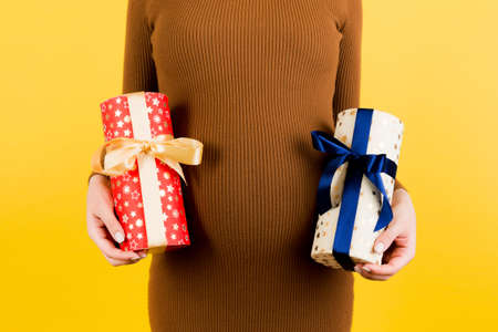 Cropped image of pregnant woman in brown dress holding two gift boxes at yellow background. Is it a boy or a girl? Waiting for twins. Pregnancy celebration. Copy space.の写真素材