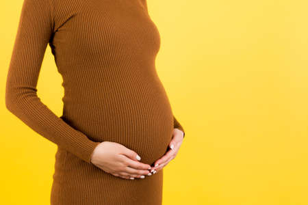 Close up of pregnant woman in brown dress holding her belly at yellow background. Motherhood concept. Copy space.の写真素材
