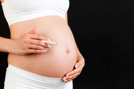 Close up of pregnant woman in white underwear with a cigarette against her belly at black background. Dangerous risk for unborn baby.の写真素材