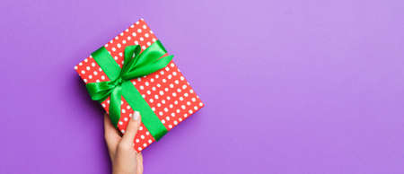 Flat lay of woman hands holding gift wrapped and decorated with bow on purple background with copy...