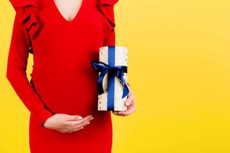 Cropped image of pregnant woman in red dress holding a gift box and touching her belly at yellow background. Expecting a baby boy. Copy space.の写真素材
