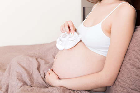 Pregnant woman With Little Baby Shoes sitting on bed in home.