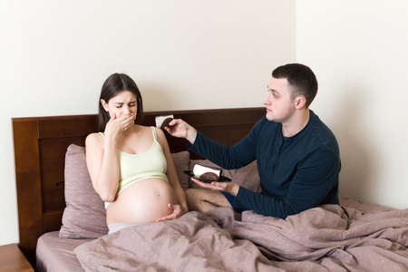 Husband offers a piece of cake to his pregnant wife but she refuses and makes stop gesture because she feels sick. Feeling bad during pregnancy concept.の写真素材
