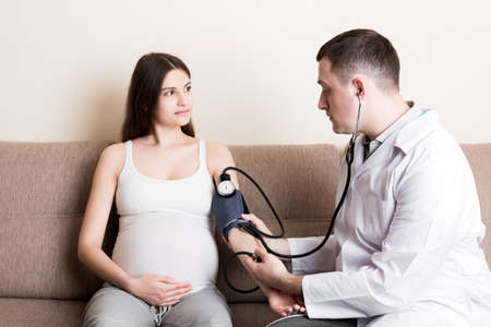 Young doctor is checking blood pressure of pregnant woman at home. High pressure problem during pregnancy concept.の写真素材