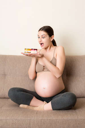 Excited pregnant woman is eating cakes with great pleasure relaxing at home. Enjoying sweet food during pregnancy concept.の写真素材