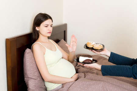 Pregnant woman relaxing in bed refuses delicious slices of cakes and makes stop gesture. Diet during pregnancy concept.の写真素材