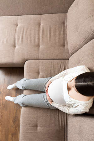 photo of cute caucasian future mom lying in light living room on sofa and holding hands on tummy.の写真素材