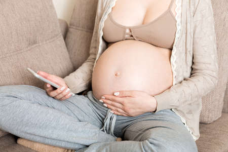 Caucasian pregnant girl using cellphone, resting on bed in home.の写真素材