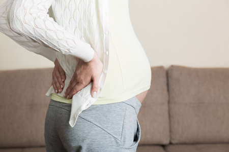 beautiful pregnant woman touching her tummy and keeping one hand on her back at home.